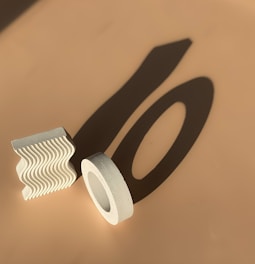 Geometric shapes, including a textured wavy block and a cylindrical ring, are casting long shadows on a beige surface under direct light.