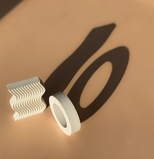 Geometric shapes, including a textured wavy block and a cylindrical ring, are casting long shadows on a beige surface under direct light.