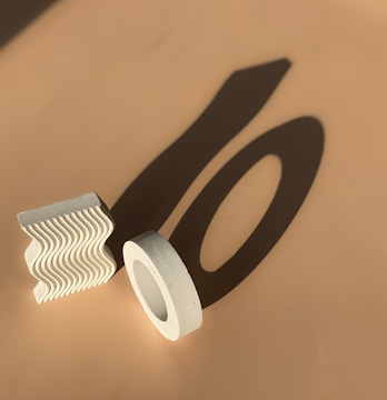 Geometric shapes, including a textured wavy block and a cylindrical ring, are casting long shadows on a beige surface under direct light.