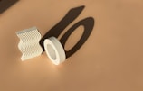 A clay sculpture set in a minimalist style featuring two geometric shapes. One shape is a wavy, vertically standing slab, the other is a circular form resembling a hollow ring. Both cast bold shadows on the smooth beige surface.