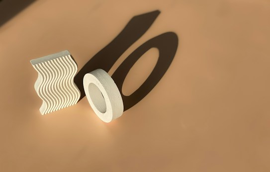 A clay sculpture set in a minimalist style featuring two geometric shapes. One shape is a wavy, vertically standing slab, the other is a circular form resembling a hollow ring. Both cast bold shadows on the smooth beige surface.