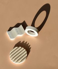 A minimalist composition with geometric shapes casting shadows on a beige surface. There are three main objects: a circular ring, a wavy pattern, and a striped circular object. The lighting creates interesting shadows, emphasizing the texture and form of each object.