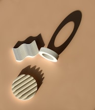 Geometric objects, including a wave-like structure, a circular object with grooves, and a ring with an extended shadow, are arranged on a flat surface. Shadows cast by these objects create interesting patterns, enhancing the visual depth.