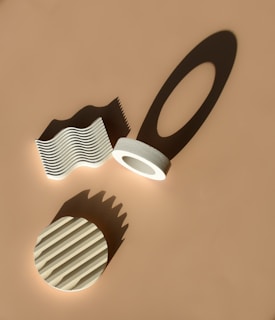 Geometric objects, including a wave-like structure, a circular object with grooves, and a ring with an extended shadow, are arranged on a flat surface. Shadows cast by these objects create interesting patterns, enhancing the visual depth.