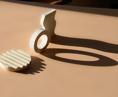 Geometric shapes made of concrete are arranged on a beige surface. The objects cast elongated shadows due to directional lighting, creating a contrast with the smooth surface.