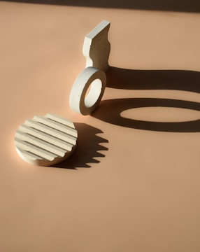 Geometric shapes are arranged on a warm, beige background, with strong lighting casting defined shadows. A circular object with textured lines sits near an upright piece that resembles a partial cylinder.