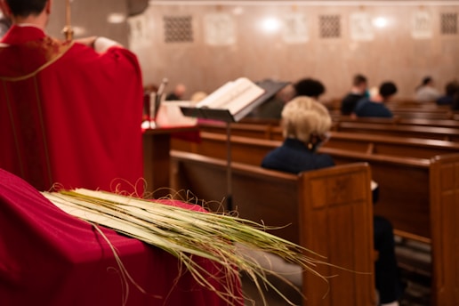 A person in a red robe stands near the front of a church, facing away from the camera. They are holding a palm frond. Behind them, several people are seated in pews, some with their heads bowed. An open book, possibly a hymnal or prayer book, is visible on a stand.