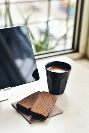 A black cup of coffee and two slices of chocolate cake are placed on a table near a window. An electronic tablet is also on the table, positioned next to the coffee and cake. The background includes a blurred outdoor view with hints of greenery.