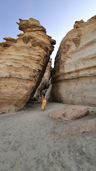 A person in a yellow dress is walking through a narrow path surrounded by towering rock formations. The surface of the rocks is smooth and layered, indicating geological weathering and erosion. The scene conveys both solitude and exploration.