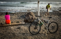 A beach scene with a person in a pink and red swimsuit sitting on a log while facing the ocean. A bicycle is leaned against a white pole in the foreground. Another person carrying a surfboard is walking towards the ocean. The beach has a rocky surface and the waves are gently crashing on the shore.