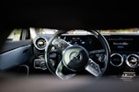 A close-up view of a car's interior focusing on the steering wheel and dashboard. The steering wheel features a prominent logo in the center, surrounded by control buttons. The dashboard has a sleek design with circular air vents, digital displays, and a portion showing text on a screen.