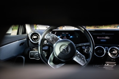 A close-up view of a car's interior focusing on the steering wheel and dashboard. The steering wheel features a prominent logo in the center, surrounded by control buttons. The dashboard has a sleek design with circular air vents, digital displays, and a portion showing text on a screen.