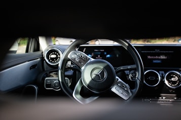 A close-up view of a car's interior focusing on the steering wheel and dashboard. The steering wheel features a prominent logo in the center, surrounded by control buttons. The dashboard has a sleek design with circular air vents, digital displays, and a portion showing text on a screen.