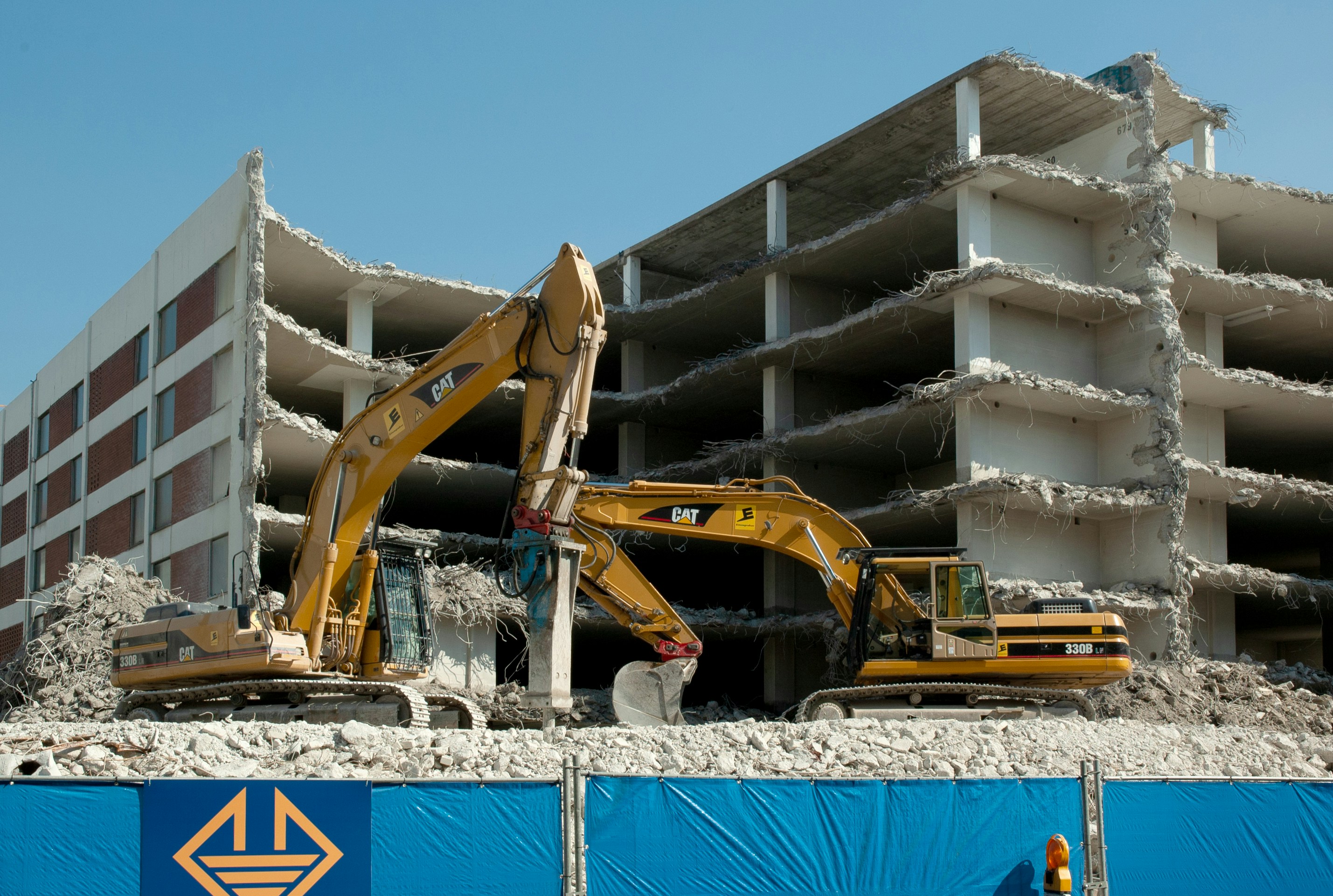 Two excavators are actively working on the demolition of a multi-story concrete building. The building is partially demolished with exposed floors and debris scattered around. A blue construction fence with a yellow logo is in the foreground.