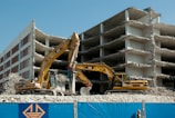 Two excavators are actively working on the demolition of a multi-story concrete building. The building is partially demolished with exposed floors and debris scattered around. A blue construction fence with a yellow logo is in the foreground.
