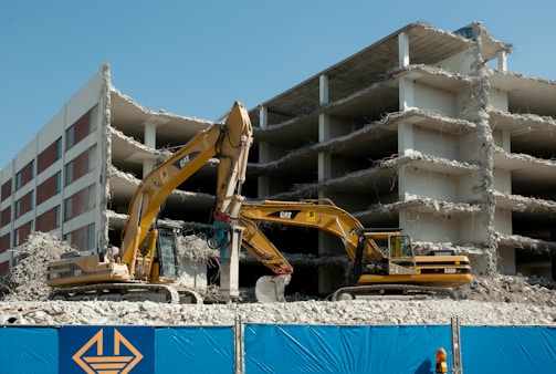 Two excavators are actively working on the demolition of a multi-story concrete building. The building is partially demolished with exposed floors and debris scattered around. A blue construction fence with a yellow logo is in the foreground.