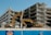 Two excavators are actively working on the demolition of a multi-story concrete building. The building is partially demolished with exposed floors and debris scattered around. A blue construction fence with a yellow logo is in the foreground.