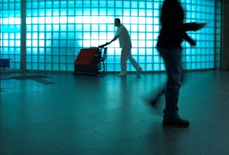 A person in a cleaning uniform is pushing a floor cleaning machine in front of a large wall made of blue-tinted glass blocks. The shadowed silhouette of another person is visible in the foreground, captured mid-step, creating a dynamic sense of motion.