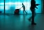 A person in a cleaning uniform is pushing a floor cleaning machine in front of a large wall made of blue-tinted glass blocks. The shadowed silhouette of another person is visible in the foreground, captured mid-step, creating a dynamic sense of motion.