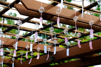 A wooden pergola adorned with numerous glass wind chimes, each with a colorful tag attached. The chimes are suspended from the beams, and the structure is surrounded by lush greenery. Sunlight filters through, casting shadows and highlighting the natural colors.