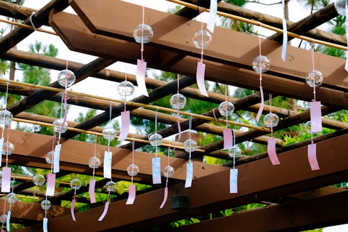 A wooden pergola adorned with numerous glass wind chimes, each with a colorful tag attached. The chimes are suspended from the beams, and the structure is surrounded by lush greenery. Sunlight filters through, casting shadows and highlighting the natural colors.
