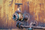 A close-up view of a rusted, industrial metal valve and piping system against a heavily corroded yellow and orange wall. The valve shows significant wear with flaking paint and visible rust, indicating age and exposure to the elements.