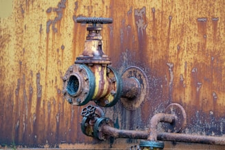 A close-up view of a rusted, industrial metal valve and piping system against a heavily corroded yellow and orange wall. The valve shows significant wear with flaking paint and visible rust, indicating age and exposure to the elements.