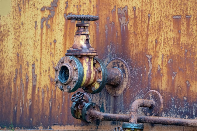 A close-up view of a rusted, industrial metal valve and piping system against a heavily corroded yellow and orange wall. The valve shows significant wear with flaking paint and visible rust, indicating age and exposure to the elements.