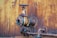 A close-up view of a rusted, industrial metal valve and piping system against a heavily corroded yellow and orange wall. The valve shows significant wear with flaking paint and visible rust, indicating age and exposure to the elements.