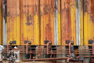 A row of industrial storage tanks with peeling and rusted yellow paint. The tanks are numbered from one to five and have corroded pipes running along the bottom. Various mechanical components and valves are visible.