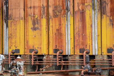 A row of industrial storage tanks with peeling and rusted yellow paint. The tanks are numbered from one to five and have corroded pipes running along the bottom. Various mechanical components and valves are visible.