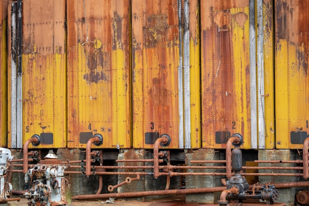 A row of industrial storage tanks with peeling and rusted yellow paint. The tanks are numbered from one to five and have corroded pipes running along the bottom. Various mechanical components and valves are visible.