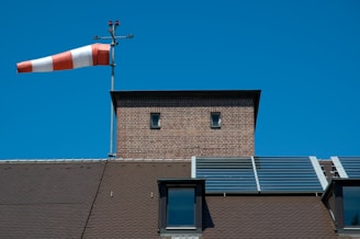 A red and white windsock is mounted on a pole atop a brick structure with a small window. The roof below is topped with solar panels and skylights, set against a clear blue sky.