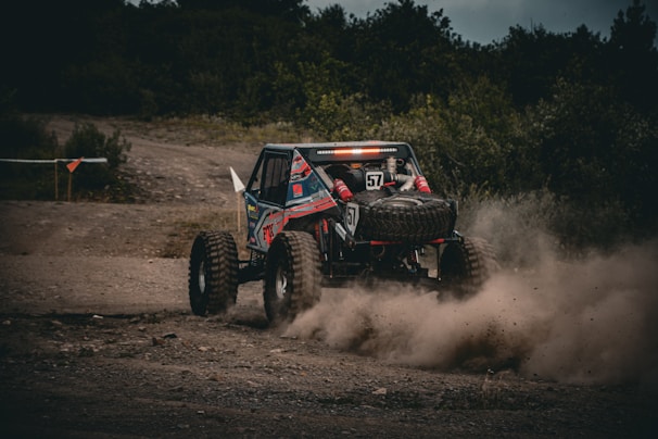 A rugged off-road vehicle speeds along a dirt path, kicking up a cloud of dust as it navigates the rough terrain. The vehicle is equipped with large tires and a roll cage, indicating it's built for challenging conditions. The background is lined with dense greenery, suggesting a remote outdoor setting.