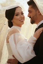 A couple embracing each other, with the woman smiling confidently while wearing an elegant white bridal gown adorned with pearls and a sheer veil. The man, dressed in a formal suit, looks lovingly at her, creating a romantic and intimate atmosphere.