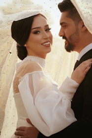 A couple embracing each other, with the woman smiling confidently while wearing an elegant white bridal gown adorned with pearls and a sheer veil. The man, dressed in a formal suit, looks lovingly at her, creating a romantic and intimate atmosphere.