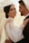 A couple embracing each other, with the woman smiling confidently while wearing an elegant white bridal gown adorned with pearls and a sheer veil. The man, dressed in a formal suit, looks lovingly at her, creating a romantic and intimate atmosphere.