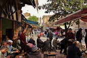 People are socializing at an outdoor cafe, surrounded by bustling street vendors and market stalls. The scene captures a lively urban environment with people engaged in conversation and enjoying beverages.