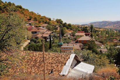 A scenic rural village nestled among hills with several small houses, mostly with red-tiled roofs. Surrounding the village are lush green trees and vegetation, and the background features rolling hills under a clear blue sky.
