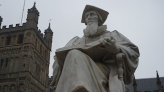 A large stone statue of a seated figure in academic robes, holding an open book, set against the backdrop of a historic building with gothic architecture.