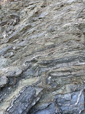 A close-up view of a textured rock surface exhibiting layers and patterns formed by geological processes. The rock displays various shades and striations, creating an intricate and natural design.