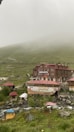 A rustic hotel and restaurant complex sits amidst a misty mountainous landscape. The buildings are constructed from wood and have red roofs. Surrounding the structures are lush green hills partially shrouded in fog. A gentle stream runs through the foreground, with some outdoor seating areas visible beside it.