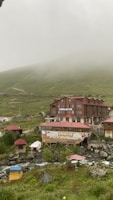 A rustic hotel and restaurant complex sits amidst a misty mountainous landscape. The buildings are constructed from wood and have red roofs. Surrounding the structures are lush green hills partially shrouded in fog. A gentle stream runs through the foreground, with some outdoor seating areas visible beside it.