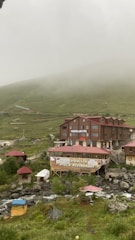 A rustic hotel and restaurant complex sits amidst a misty mountainous landscape. The buildings are constructed from wood and have red roofs. Surrounding the structures are lush green hills partially shrouded in fog. A gentle stream runs through the foreground, with some outdoor seating areas visible beside it.