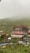 A rustic hotel and restaurant complex sits amidst a misty mountainous landscape. The buildings are constructed from wood and have red roofs. Surrounding the structures are lush green hills partially shrouded in fog. A gentle stream runs through the foreground, with some outdoor seating areas visible beside it.