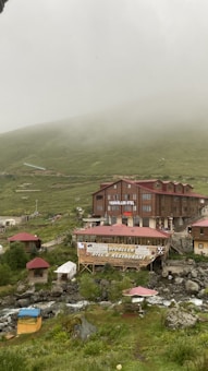 A rustic hotel and restaurant complex sits amidst a misty mountainous landscape. The buildings are constructed from wood and have red roofs. Surrounding the structures are lush green hills partially shrouded in fog. A gentle stream runs through the foreground, with some outdoor seating areas visible beside it.