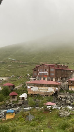 A rustic hotel and restaurant complex sits amidst a misty mountainous landscape. The buildings are constructed from wood and have red roofs. Surrounding the structures are lush green hills partially shrouded in fog. A gentle stream runs through the foreground, with some outdoor seating areas visible beside it.