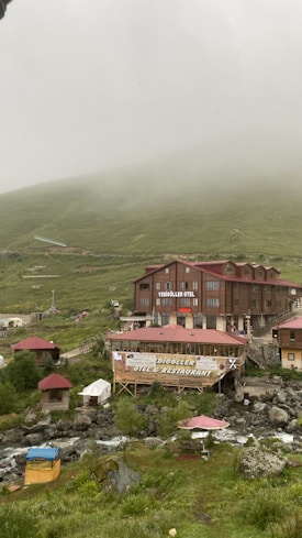 A rustic hotel and restaurant complex sits amidst a misty mountainous landscape. The buildings are constructed from wood and have red roofs. Surrounding the structures are lush green hills partially shrouded in fog. A gentle stream runs through the foreground, with some outdoor seating areas visible beside it.