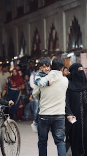 A bustling street scene depicts a man carrying a crying child in his arms, flanked by a woman dressed in black attire. The crowd in the background suggests a busy marketplace or shopping area. A bicycle is partially visible at the edge of the frame.