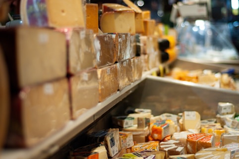 A selection of various cheeses displayed on shelves in a store. The cheeses are wrapped and stacked neatly, with some being oversized blocks and others in smaller packages. The setting appears to be a market or deli with a variety of cheese textures and colors visible.